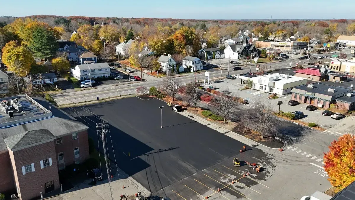 Aerial view of a town square with a newly paved parking lot, surrounded by buildings and trees in autumn colors.
