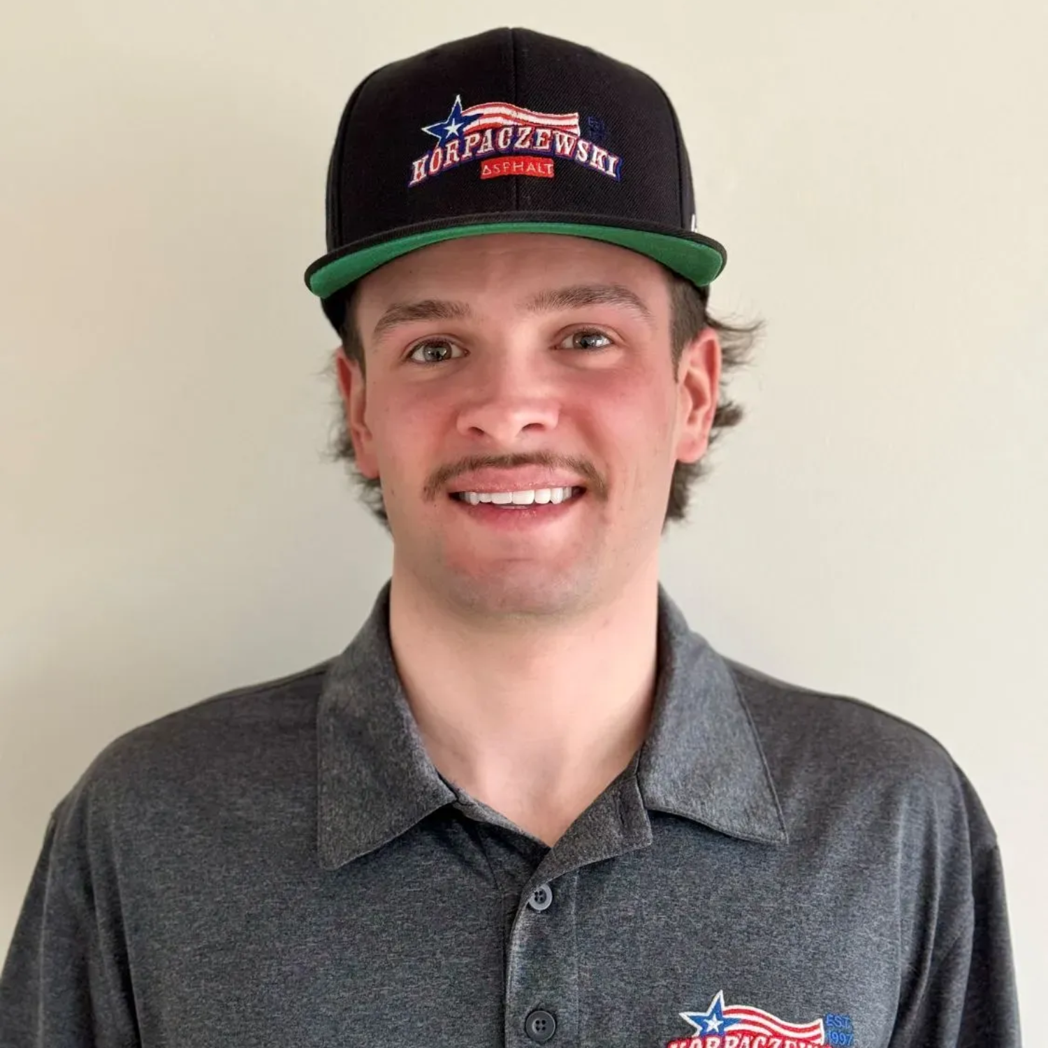 Man smiling, wearing a black cap with a logo, and a gray polo shirt.