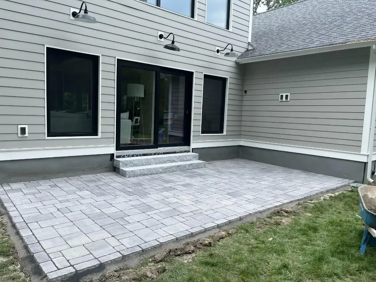 Newly installed brick patio outside a house with sliding glass door. Gray siding, steps, and grass.