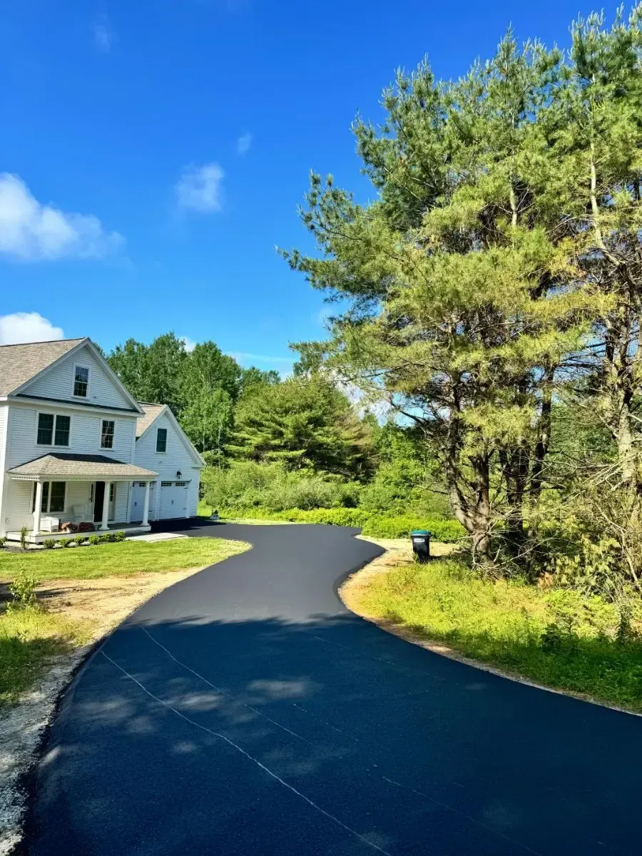 Asphalt driveway leading to a gray house with a two-car garage under a bright blue sky. Green trees surround.