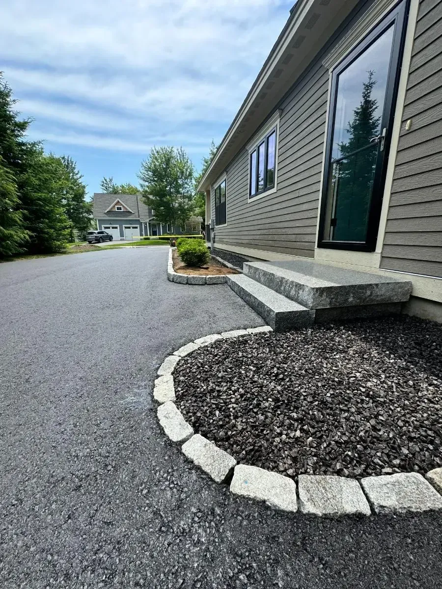 Asphalt driveway leads to house with steps and flower bed, blue sky overhead.