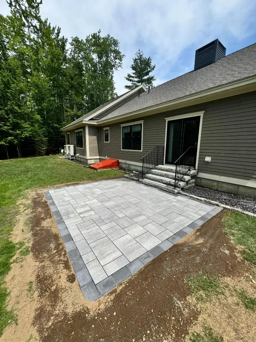 Patio paved with gray bricks bordered by darker bricks, next to a house with a door, steps, and handrail.