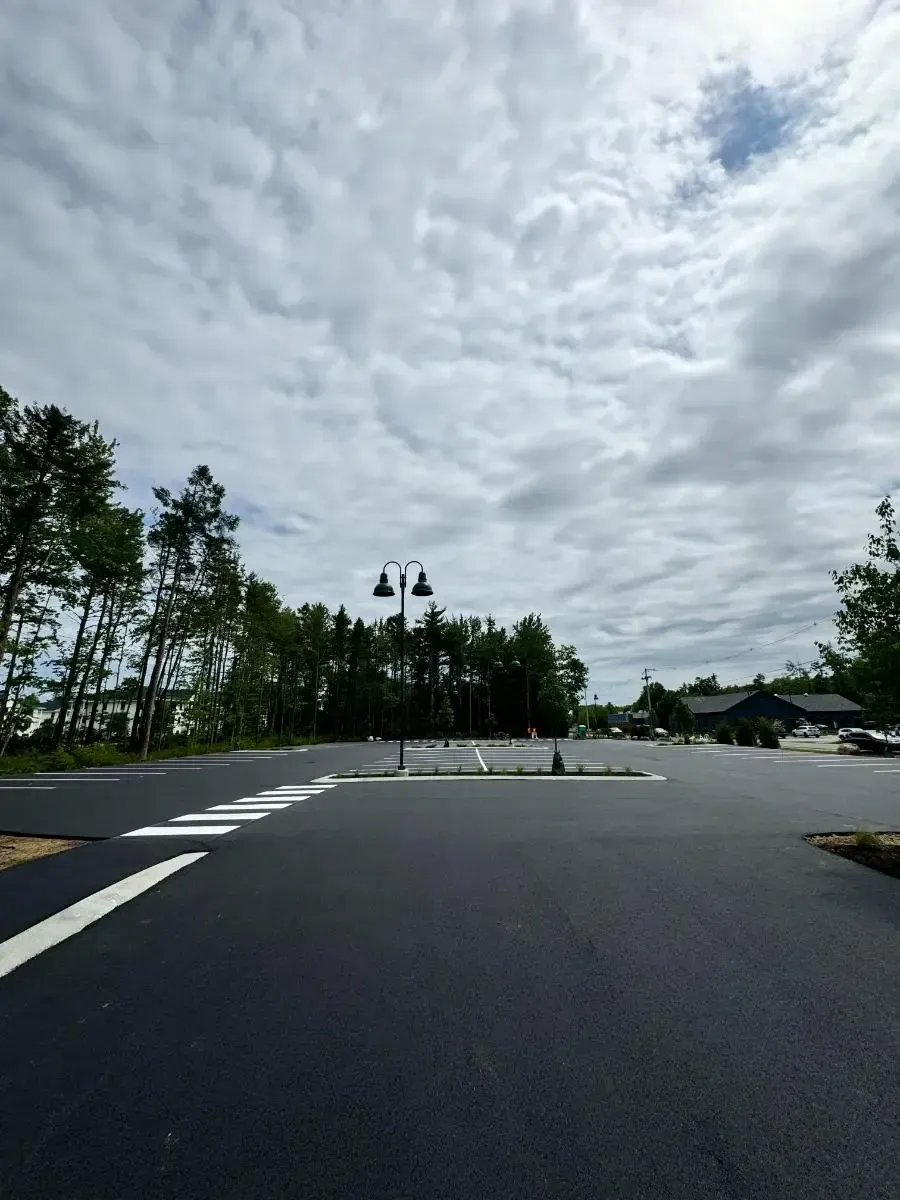 Black paved parking lot under a cloudy sky, trees in the background. A decorative street lamp is in the center.