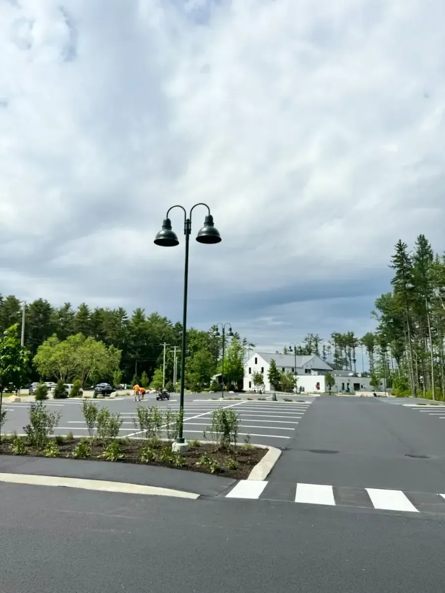 Empty parking lot with street lamp; white buildings and trees in the background under a cloudy sky.