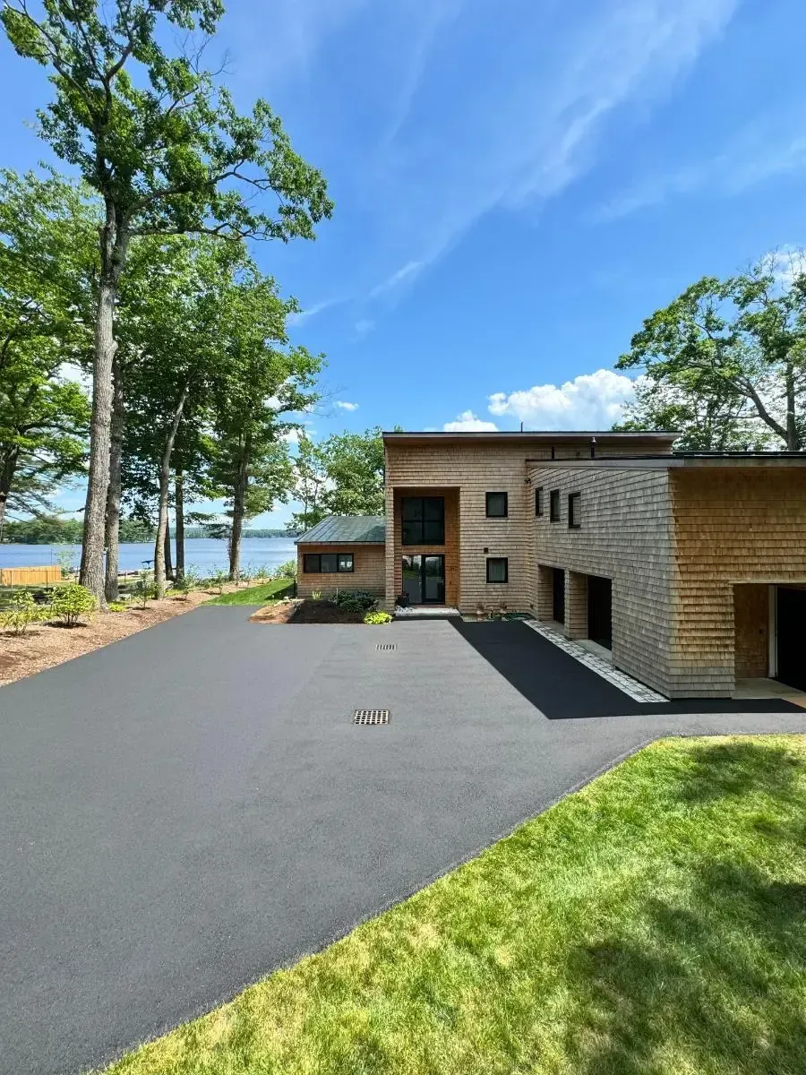 Modern house with wooden accents and asphalt driveway, on a lake under a sunny sky.