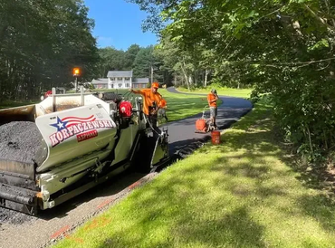 Road crew paving a road with asphalt. Workers in orange vests guide a machine on a tree-lined street.