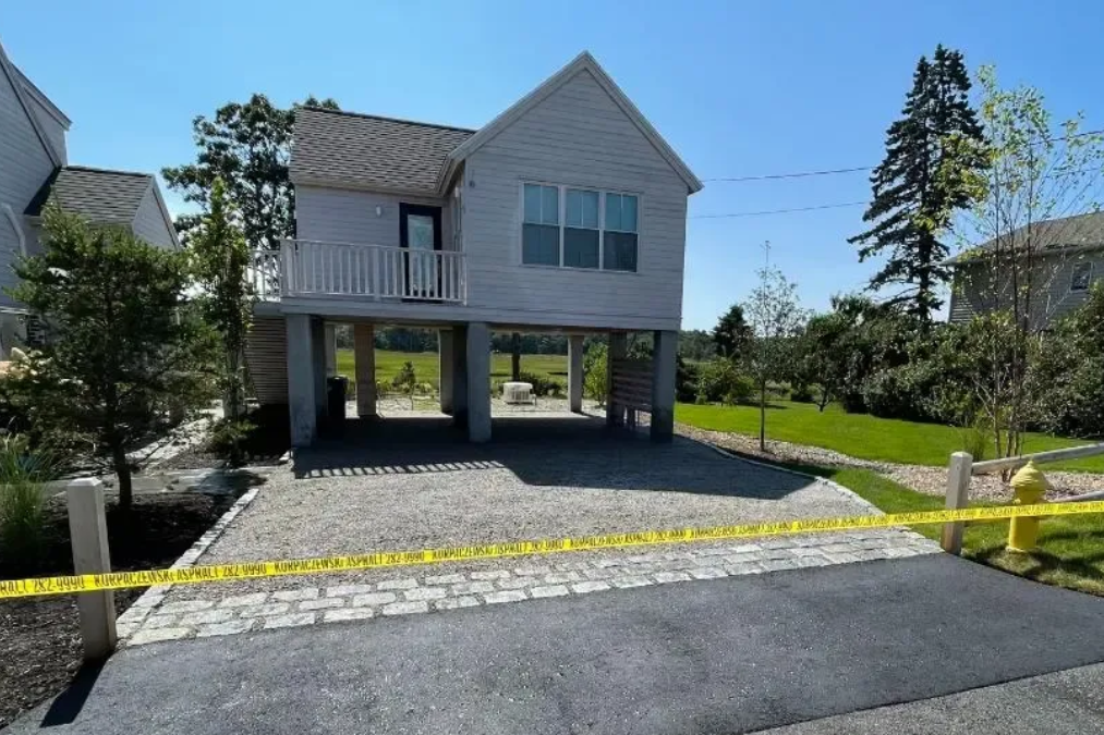Small house raised on pillars with gravel driveway, yellow caution tape, and green lawn on a sunny day.