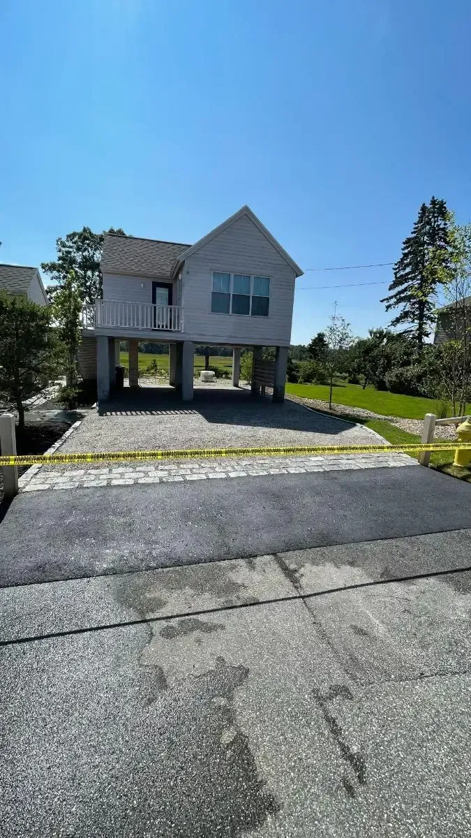 Elevated gray house on stilts with gravel and new asphalt driveway, yellow caution tape. Bright blue sky.