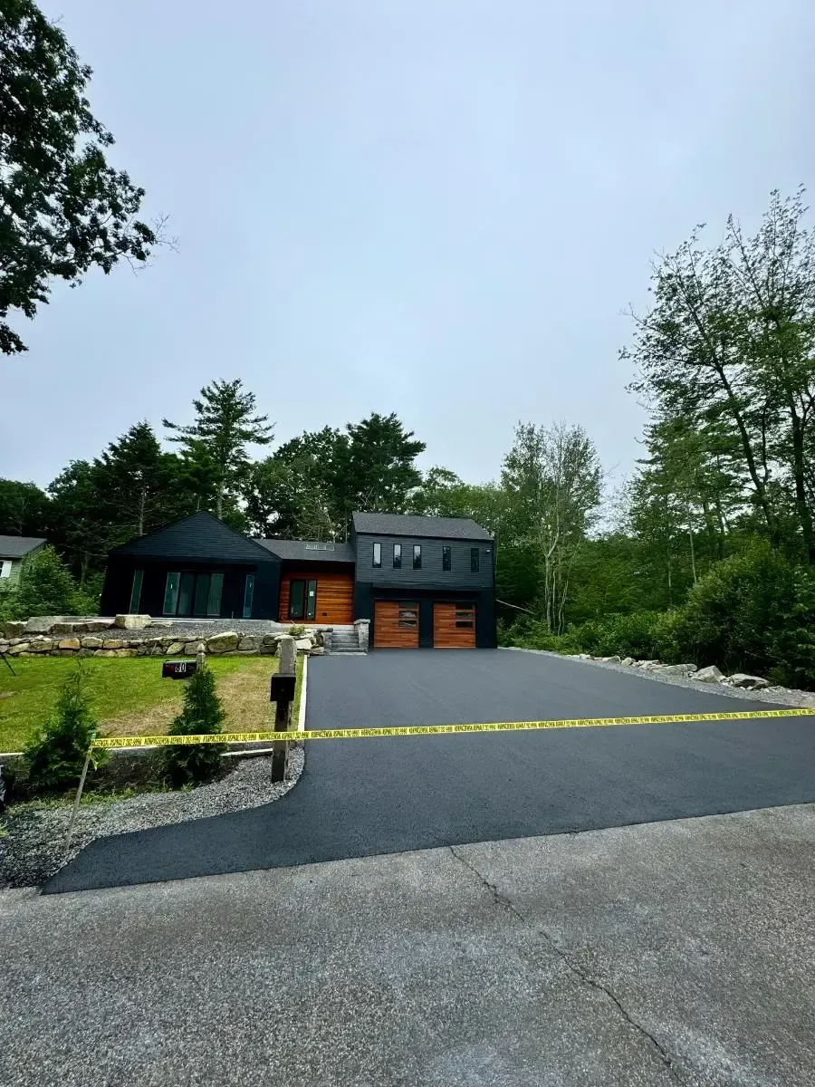 Modern black house with wood garage doors, fresh asphalt driveway, and caution tape.