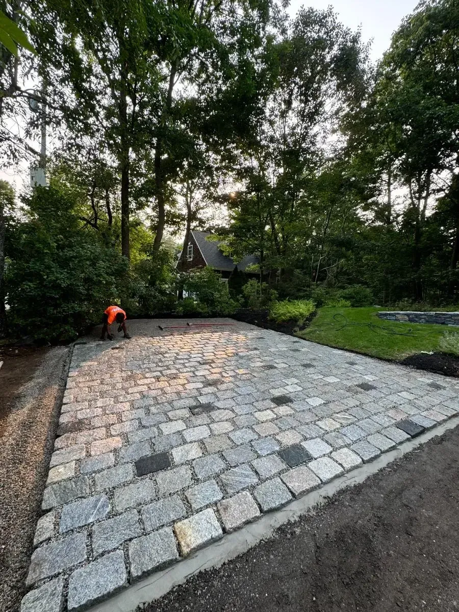 Cobblestone patio under construction, person working. Trees and a house in the background.