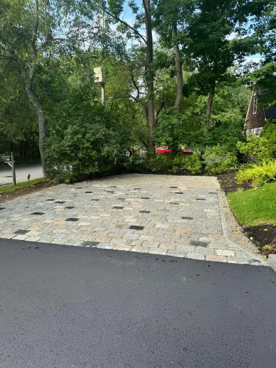 Asphalt driveway leads to a stone and brick parking area with trees in the background.