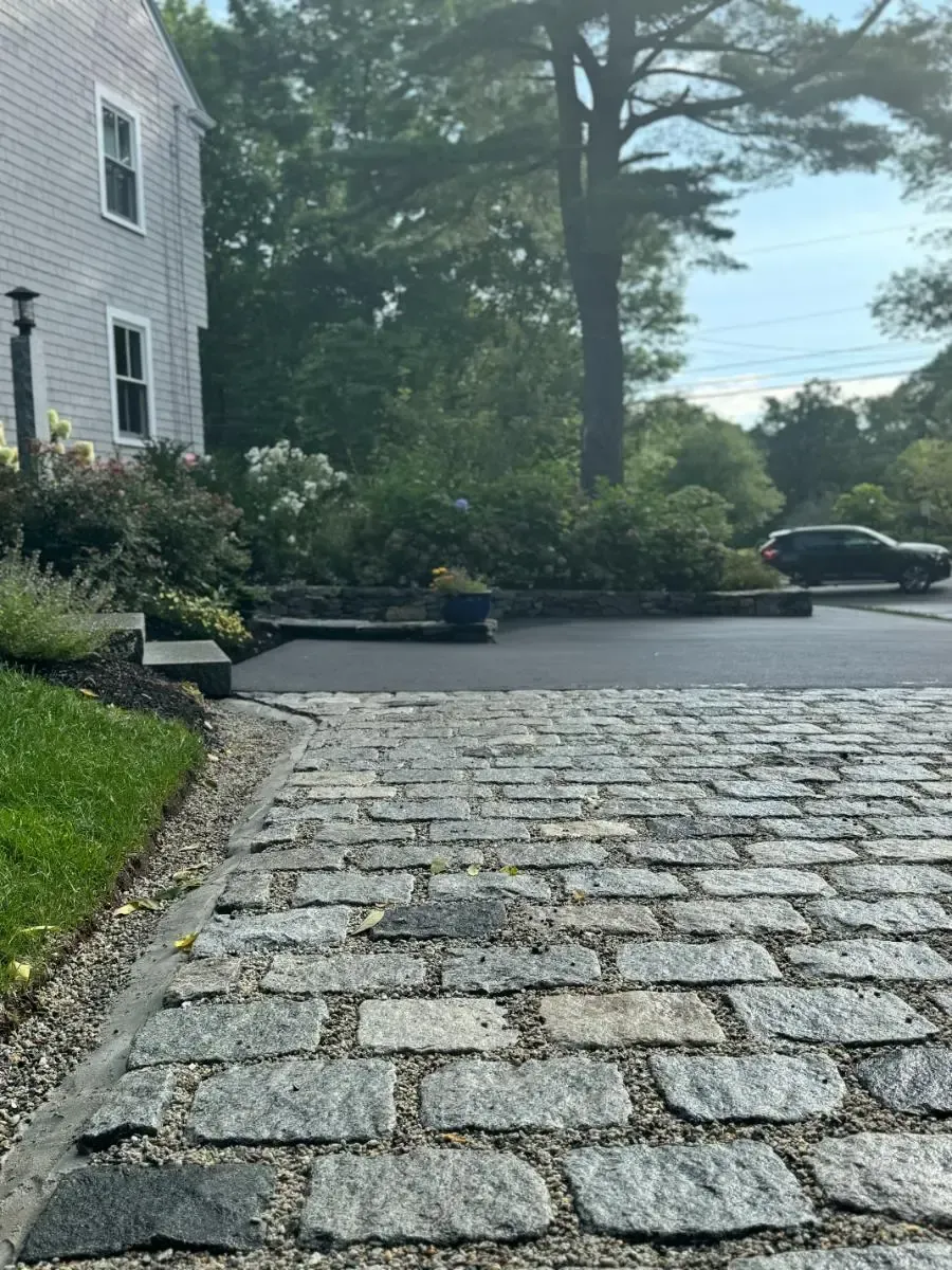 Cobblestone driveway leading to a house with asphalt and landscaping; a car is in the background.