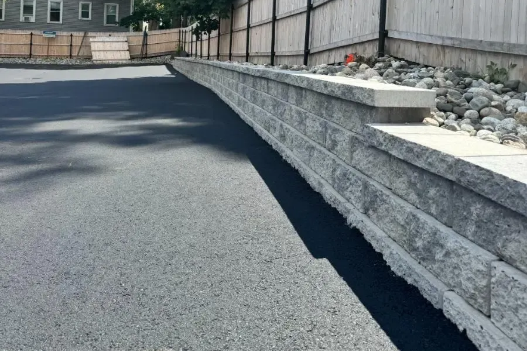 Asphalt driveway next to a gray stone retaining wall. A wooden fence is in the background.