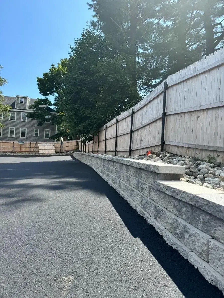 Asphalt driveway slopes up to a retaining wall and wooden fence. Bright sunlight.