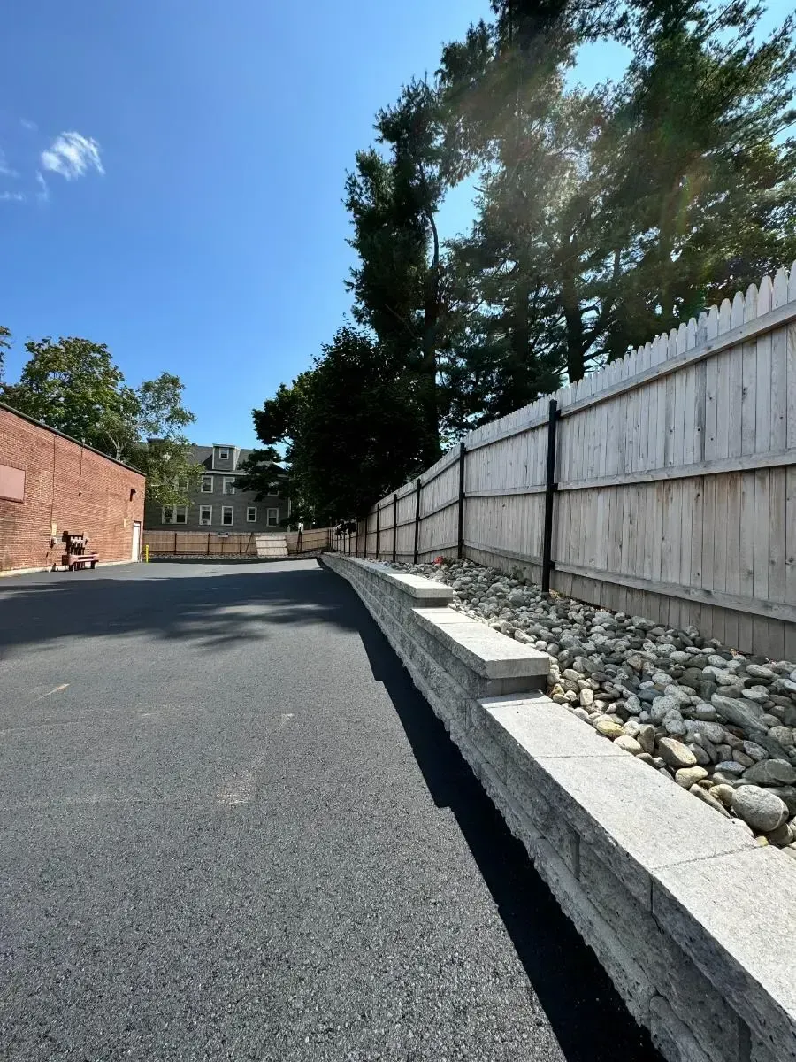 Asphalt parking area with a stone retaining wall and wooden fence against a bright blue sky.