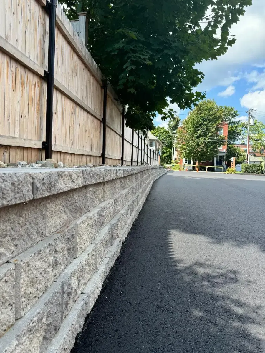 A paved road curves past a retaining wall, with a wooden fence and trees in the background under a cloudy sky.