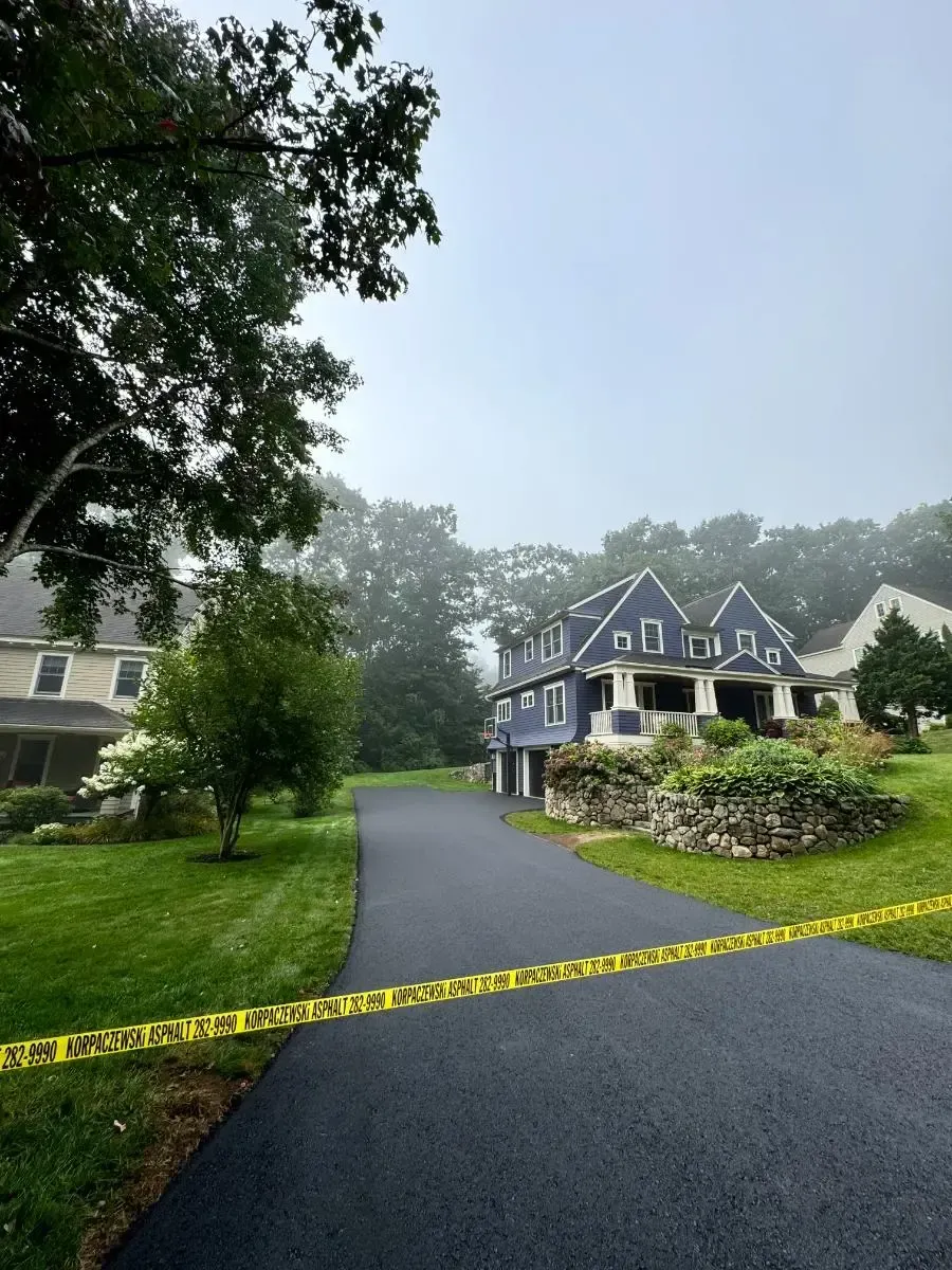 Asphalt driveway leading to a gray house with a two-car garage under a bright blue sky. Green trees surround.