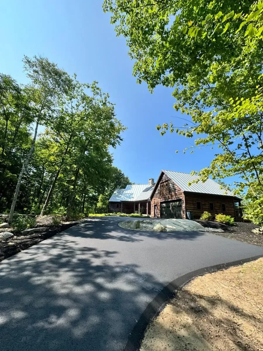 Asphalt driveway leading to a brown building with a metal roof surrounded by trees under a blue sky.