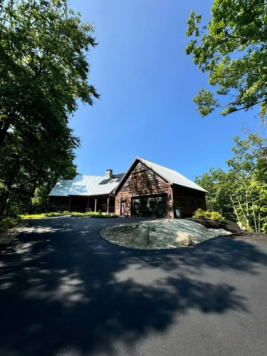 Rustic wooden building with metal roof; dark asphalt driveway leads to the entrance framed by trees, bright blue sky.