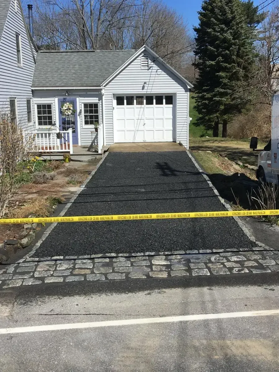 Newly paved asphalt driveway leading to a white garage, bordered by cobblestones, with a yellow caution tape in the foreground.