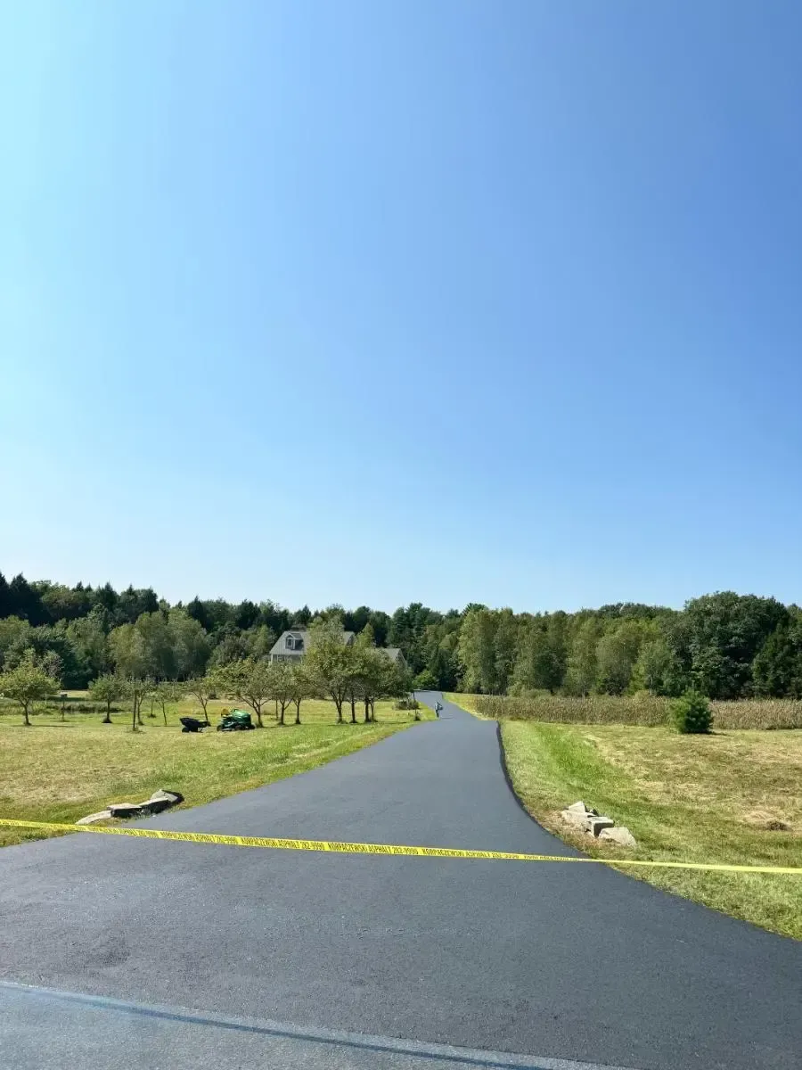 A newly paved asphalt road leads toward a house surrounded by trees under a clear blue sky.