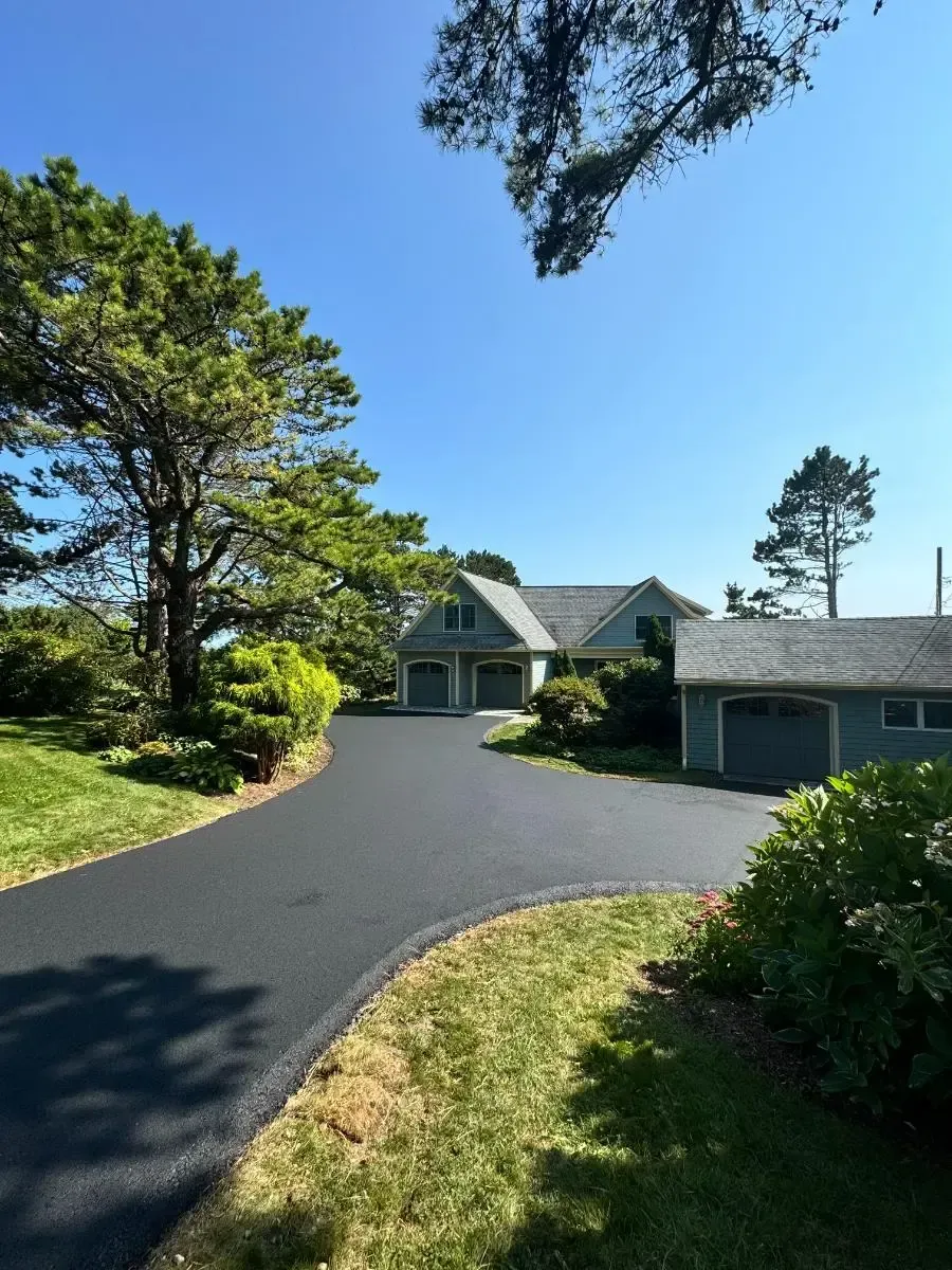 Asphalt driveway leading to a gray house with a two-car garage under a bright blue sky. Green trees surround.