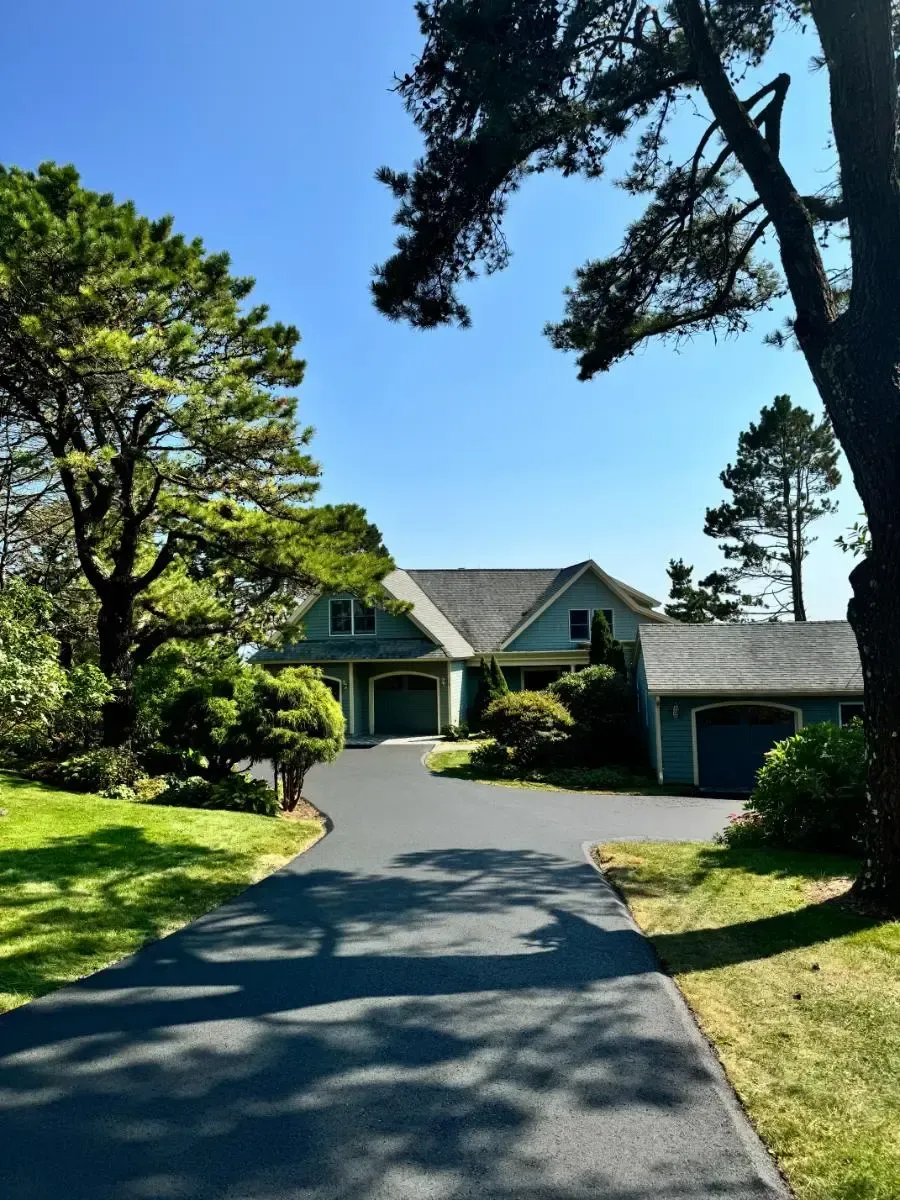 House with teal siding, dark driveway, and green lawn under a blue sky, framed by trees.