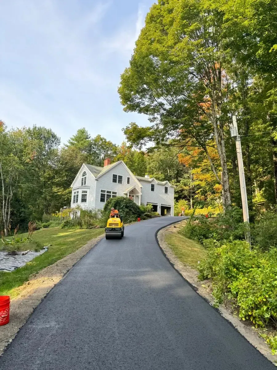 Asphalt driveway leading to a white house, with a small roller compactor; trees surround the home.