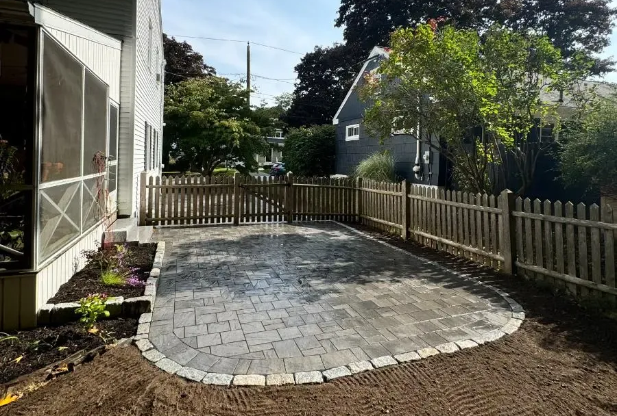 Backyard patio with gray pavers and a wooden fence, surrounded by dirt and grass.