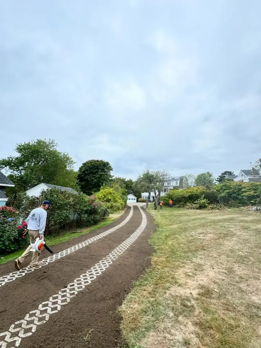 Person using a leaf blower on a decorative gravel driveway, cloudy sky overhead.