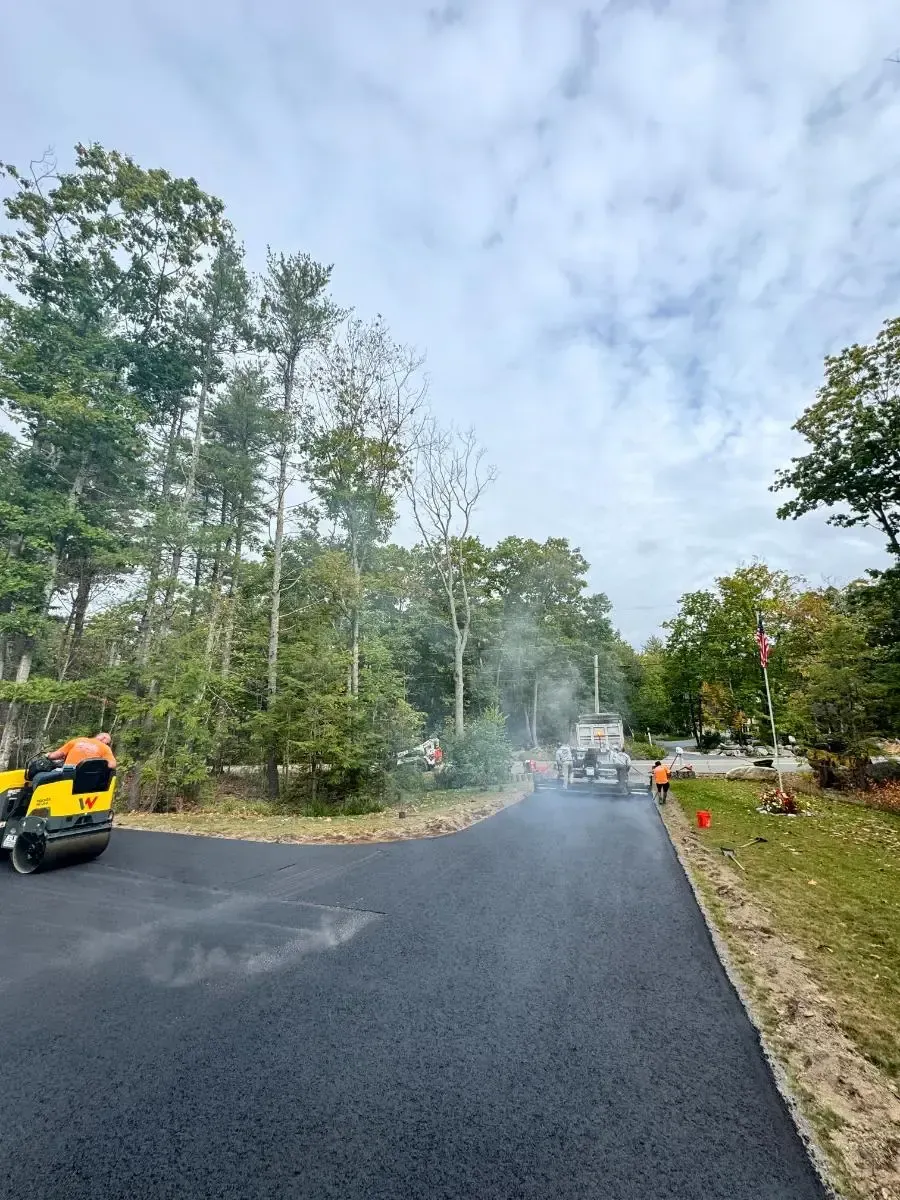 Asphalt paving in progress: machine laying blacktop on a dirt road, worker guiding. Rolling machine nearby.