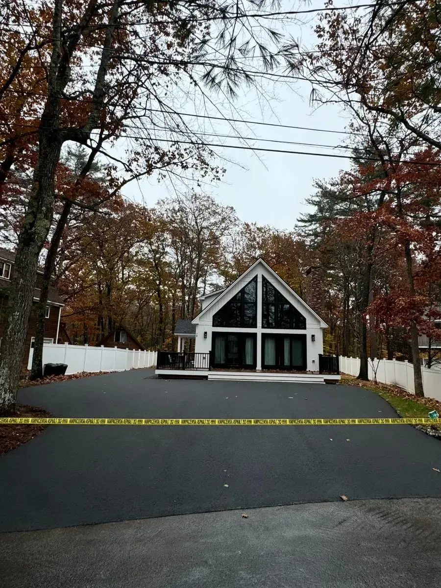 Asphalt paving roadwork. Paving machine, workers, orange cones, grass, trees, and residential setting. Sunny day.
