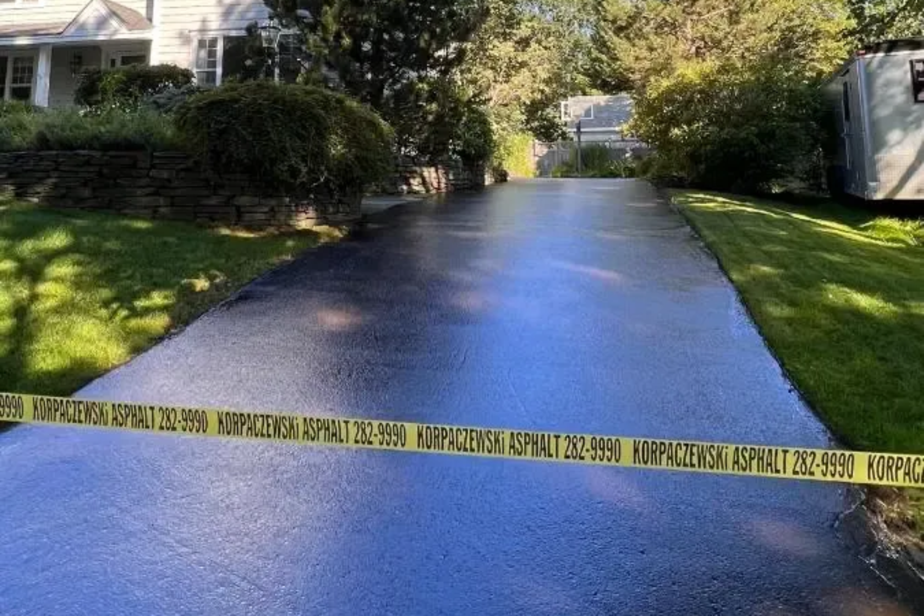 Freshly sealed blacktop driveway with yellow caution tape, surrounded by green grass and trees.