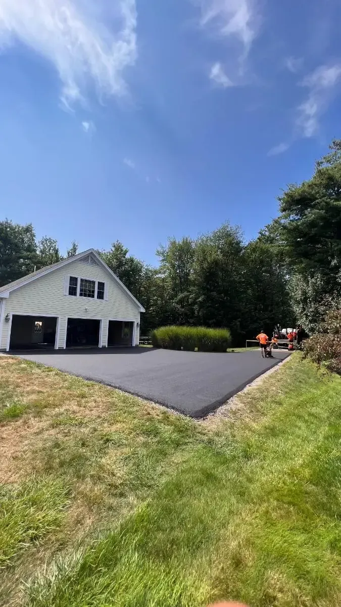 Asphalt driveway leading to a white two-car garage under a blue sky, surrounded by green grass and trees.