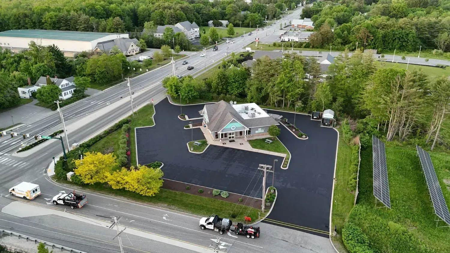 Aerial view of a building with a dark parking lot next to a road, surrounded by greenery and trees.