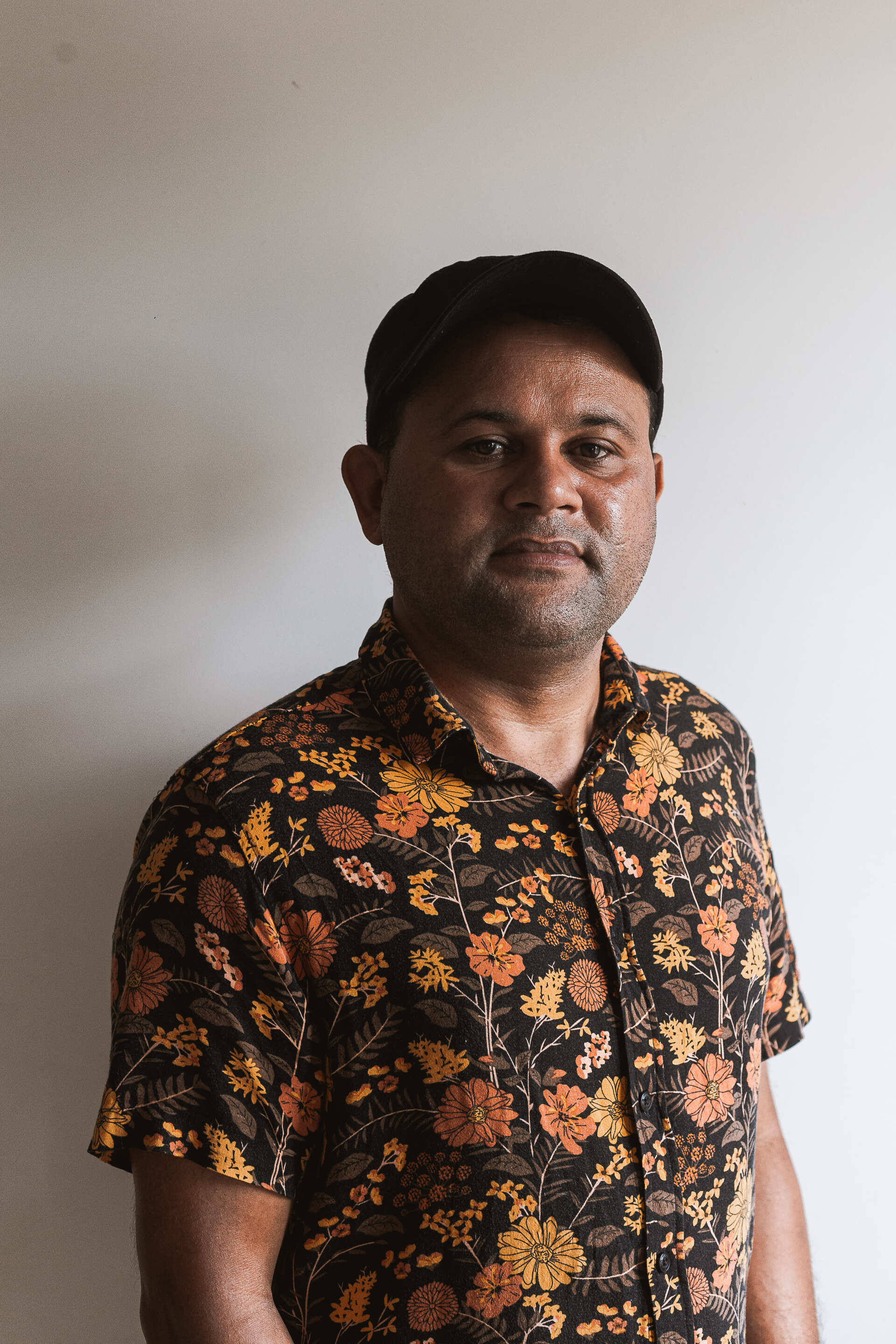 Um homem de chapéu e camisa floral está parado em frente a uma parede branca.