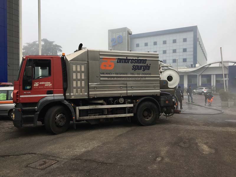 Un camion rosso e argento per le acque reflue parcheggiato vicino a un edificio con una colonna di vapore, probabilmente impegnato nelle pulizie.