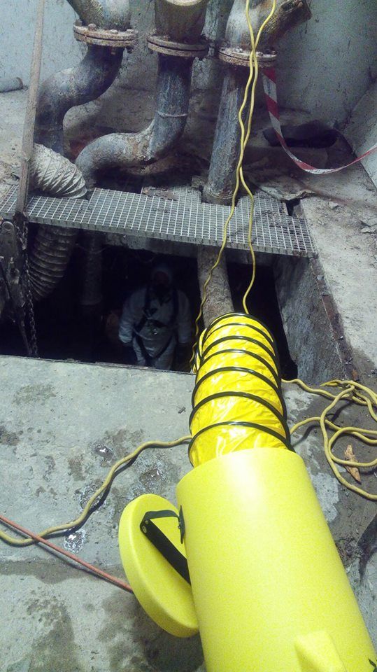 Lavoratore in uno spazio confinato, che utilizza un tubo di ventilazione giallo. Tubi e grate sono visibili.