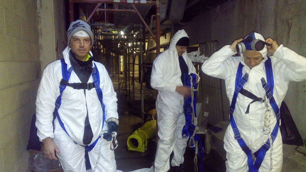 Tre persone con tute protettive bianche e imbracature, impegnate nei preparativi o al lavoro in un cantiere edile.