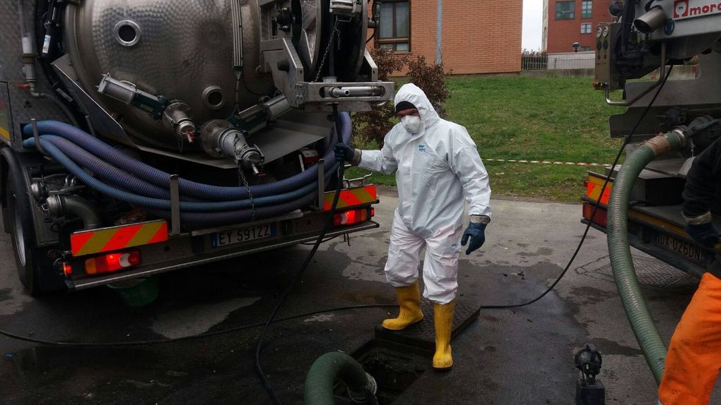 Persona in tuta protettiva al lavoro su un'autocisterna per lo scarico delle acque reflue, mentre collega i tubi flessibili. All'aperto, in un ambiente industriale.