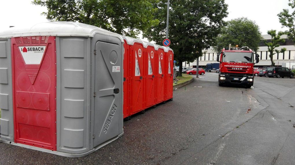 Fila di bagni chimici, rossi e grigi, lungo una strada con un camion dei pompieri sullo sfondo.