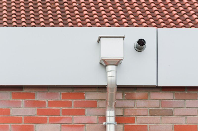 Dettagli dell'edificio esterno: grondaia e pluviale in metallo su muro di mattoni, telecamera di sicurezza e tetto in tegole rosse.