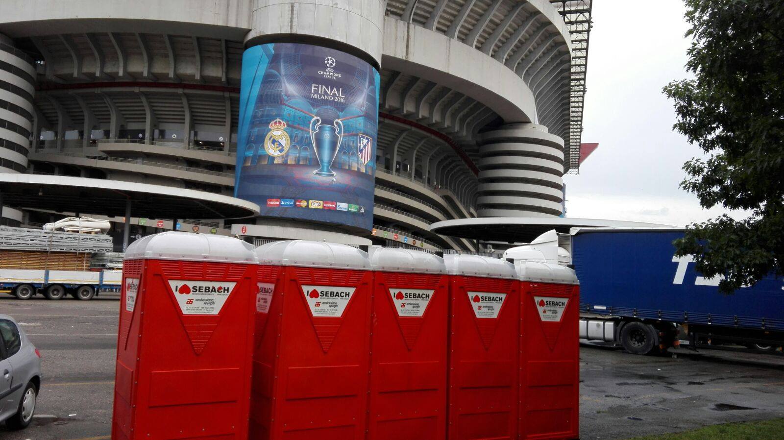 Bagni chimici rossi davanti a uno stadio con uno striscione della Champions League, un camion parcheggiato e un'auto.