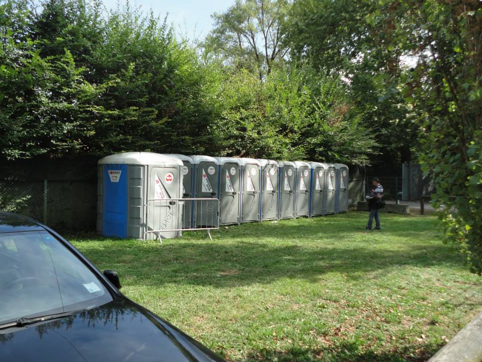 Fila di bagni chimici sull'erba, accanto a una recinzione e a dei cespugli. Una persona è in piedi lì vicino.