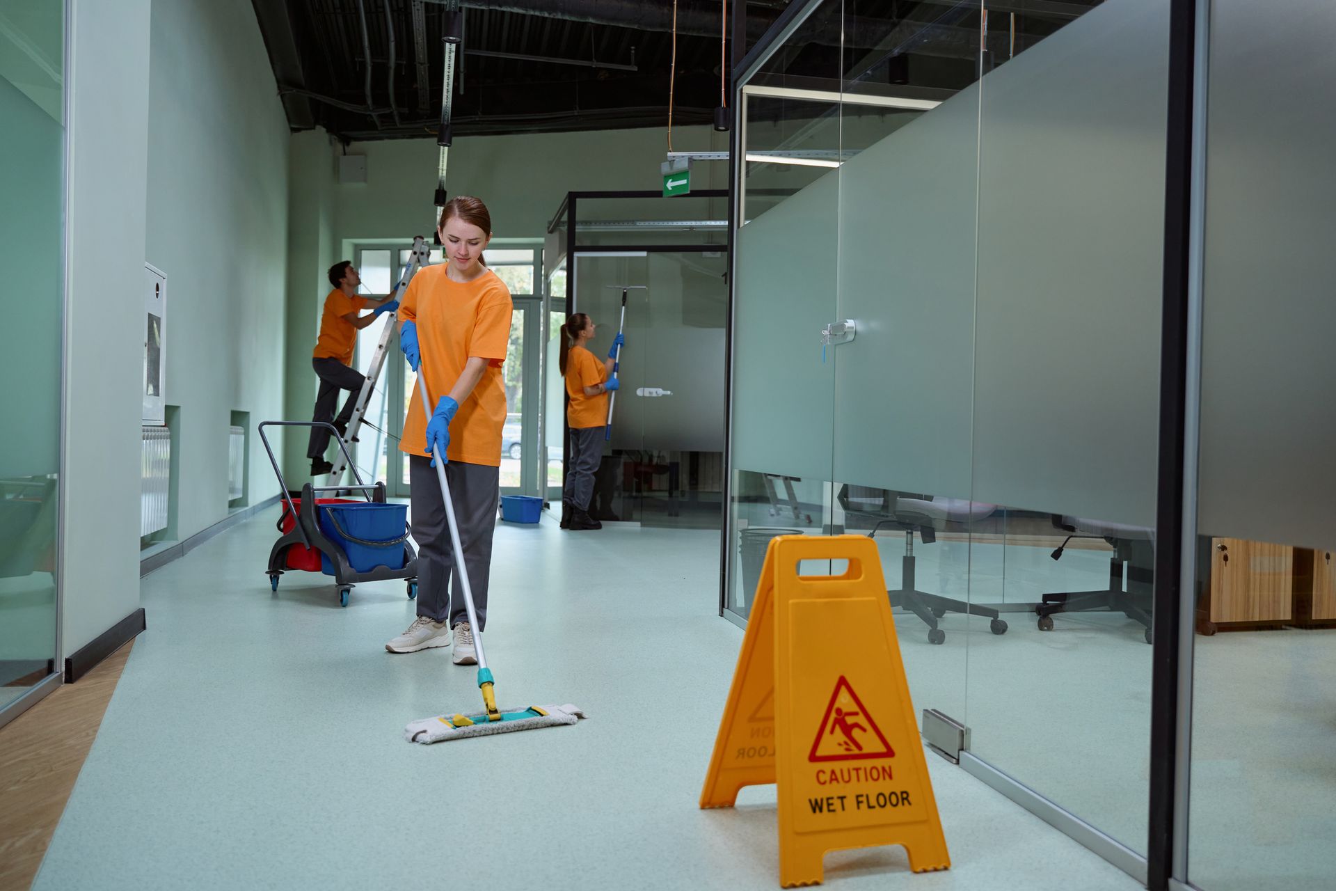 A woman is mopping the floor of an office building next to a caution sign.