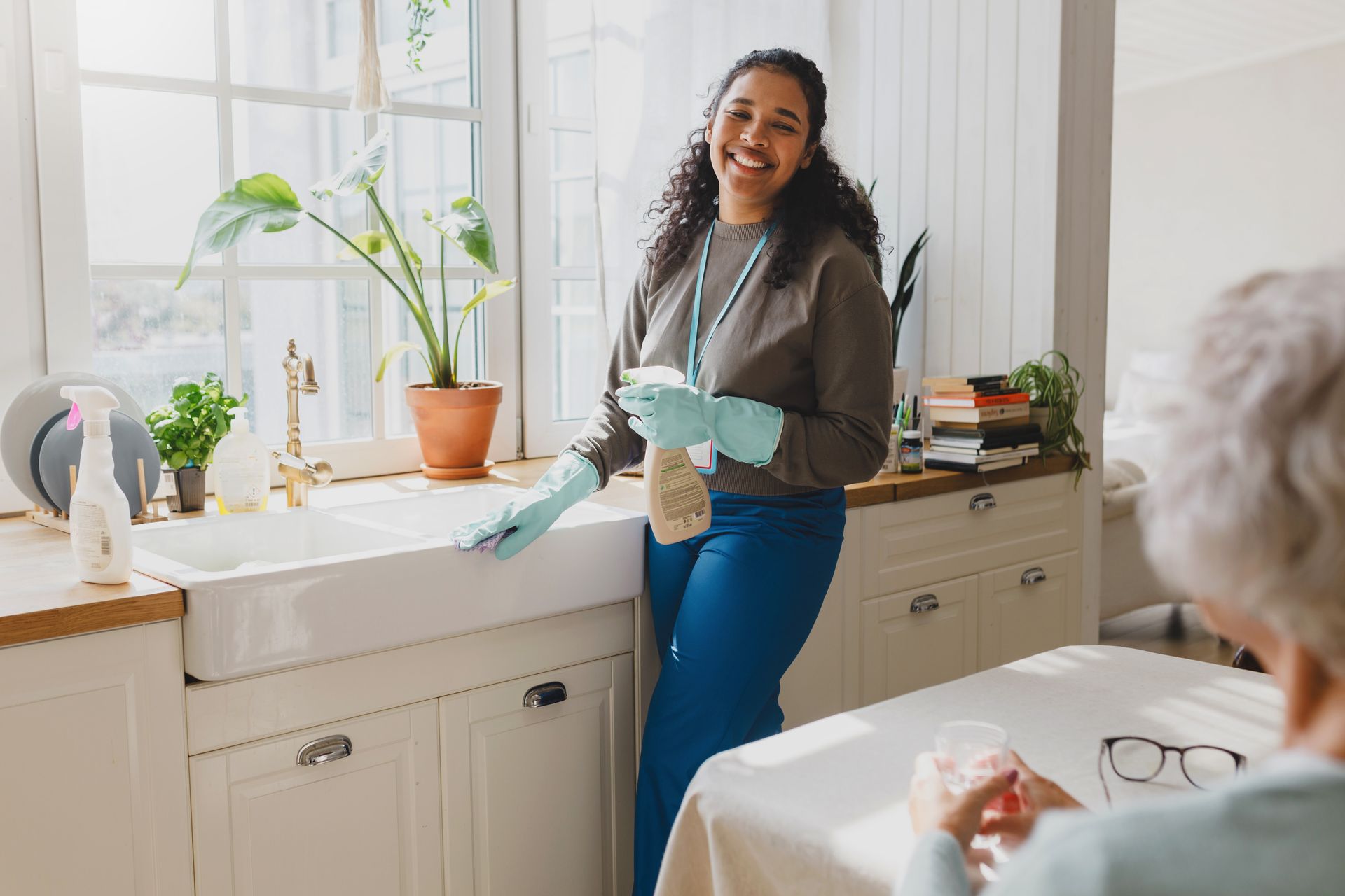 A woman is cleaning a kitchen sink while an elderly woman sits at a table.
