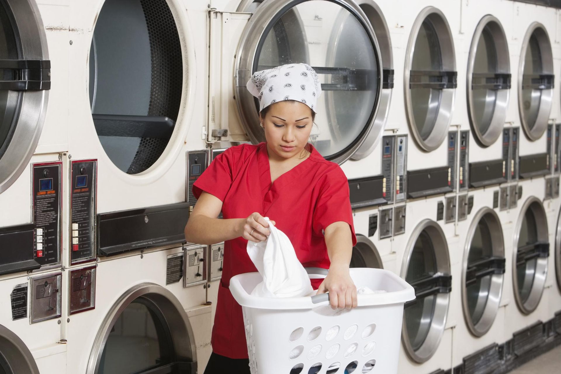 A woman in a red scrub top is holding a laundry basket in a laundromat