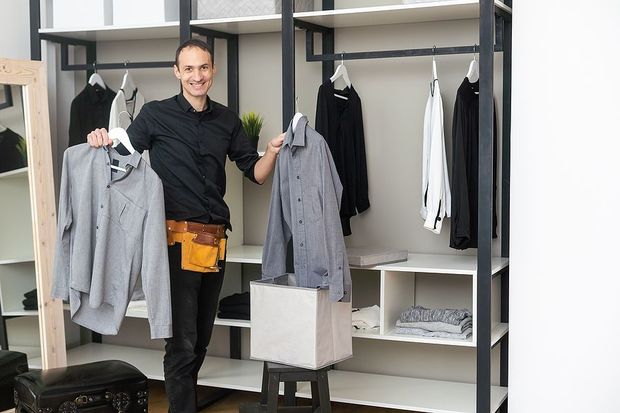 A man is holding a sweater and a shirt in a closet.