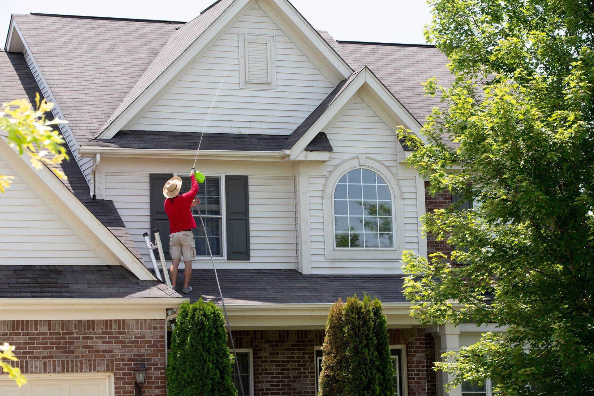 A man is standing on a ladder cleaning the roof of a house.