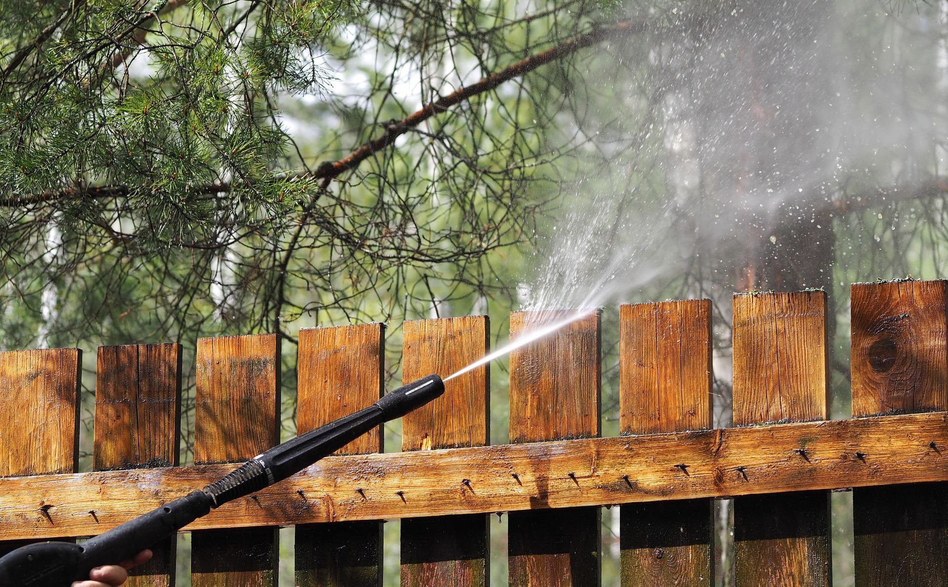 A person is using a high pressure washer to clean a wooden fence.